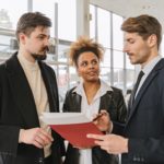 The Importance of Understanding Tenants Needs in Commercial Real Estate Investments. Image of three people standing in a building. One person is talking and holding a red folder with paperwork while the other two listen and look at him.